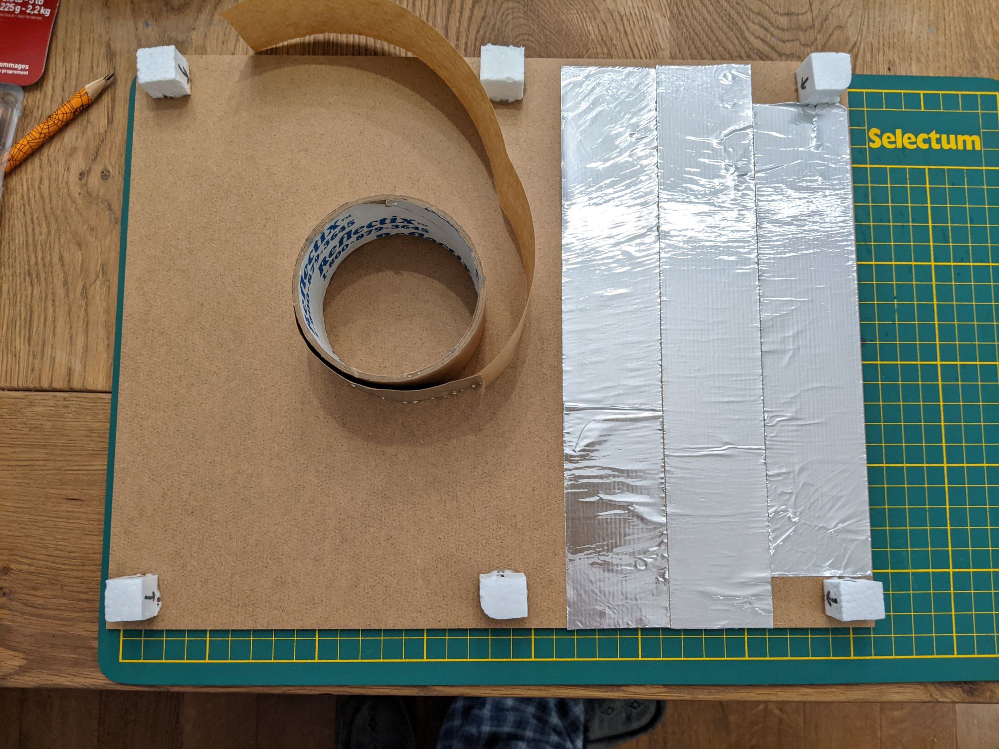 Backing board with styrofoam spacers and foil tape on a cutting mat. There is a roll of foil tape on top.